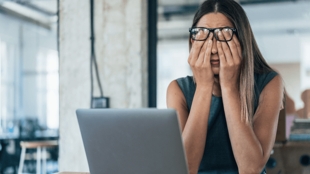 Mulher estressada cobrindo o rosto diante do computador, simbolizando assédio disfarçado de brincadeira no ambiente de trabalho